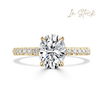 A yellow gold oval solitaire engagement ring featuring a large oval-cut center diamond and a pavé-set diamond band, shown on a clean white background with an 'In Stock' text overlay in the top right.