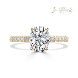 A yellow gold oval solitaire engagement ring featuring a large oval-cut center diamond and a pavé-set diamond band, shown on a clean white background with an 'In Stock' text overlay in the top right.